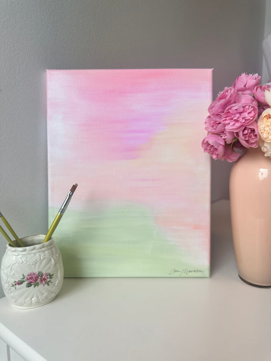 An abstract pastel canvas painting with pink, peach, cream, and mint green watercolor-like washes, displayed on a white tabletop next to a vase of pink flowers and a small floral paint holder with brushes.