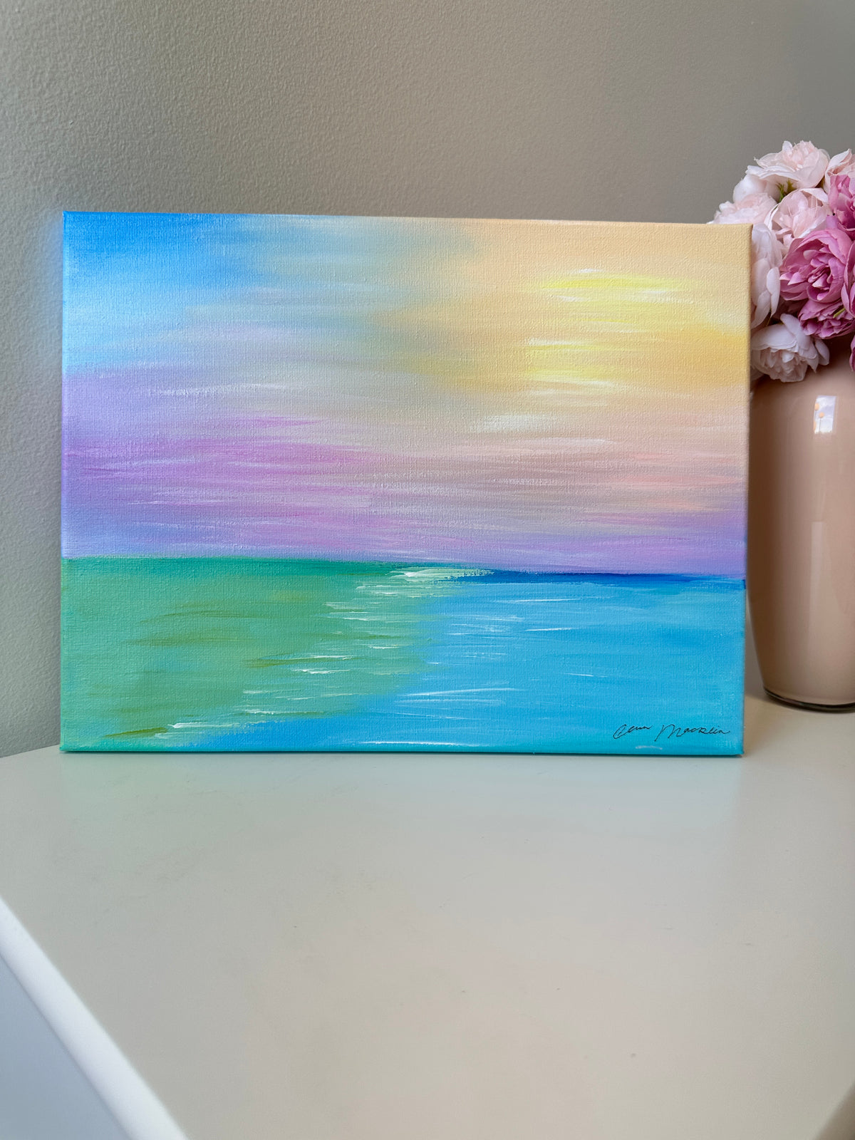 A rectangle pastel landscape painting of an ocean sunset, displayed on a white tabletop next to a pink flower vase. The painting features a soft sky gradient of blue, yellow, pink, and purple over a turquoise sea.