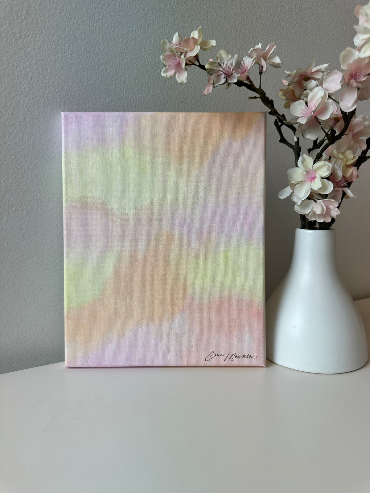 A square canvas painting with a soft pastel watercolor design in shades of pink, yellow, and peach, displayed on a white surface next to a white vase with pink blossoms against a gray wall.