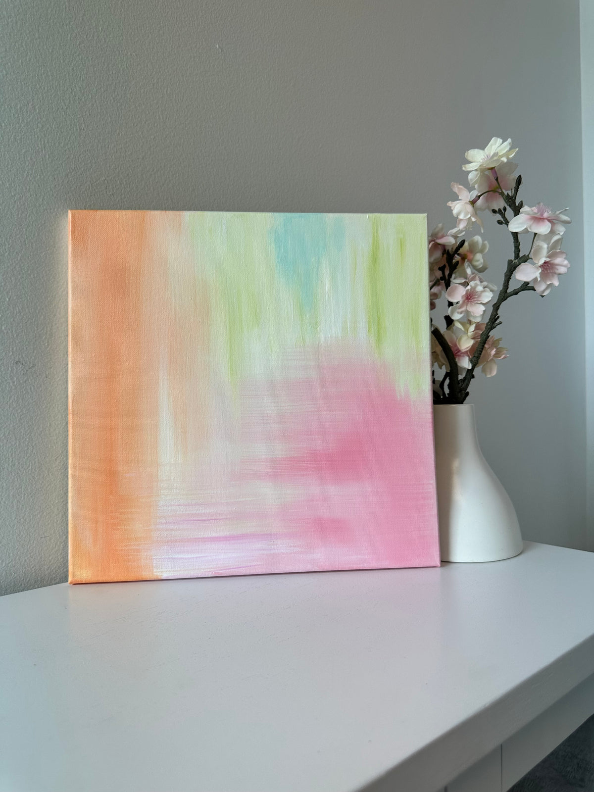 An abstract pastel canvas painting with soft gradients of pink, peach, mint green, and light blue, displayed on a white table next to a white vase with pink blossoms against a gray wall.