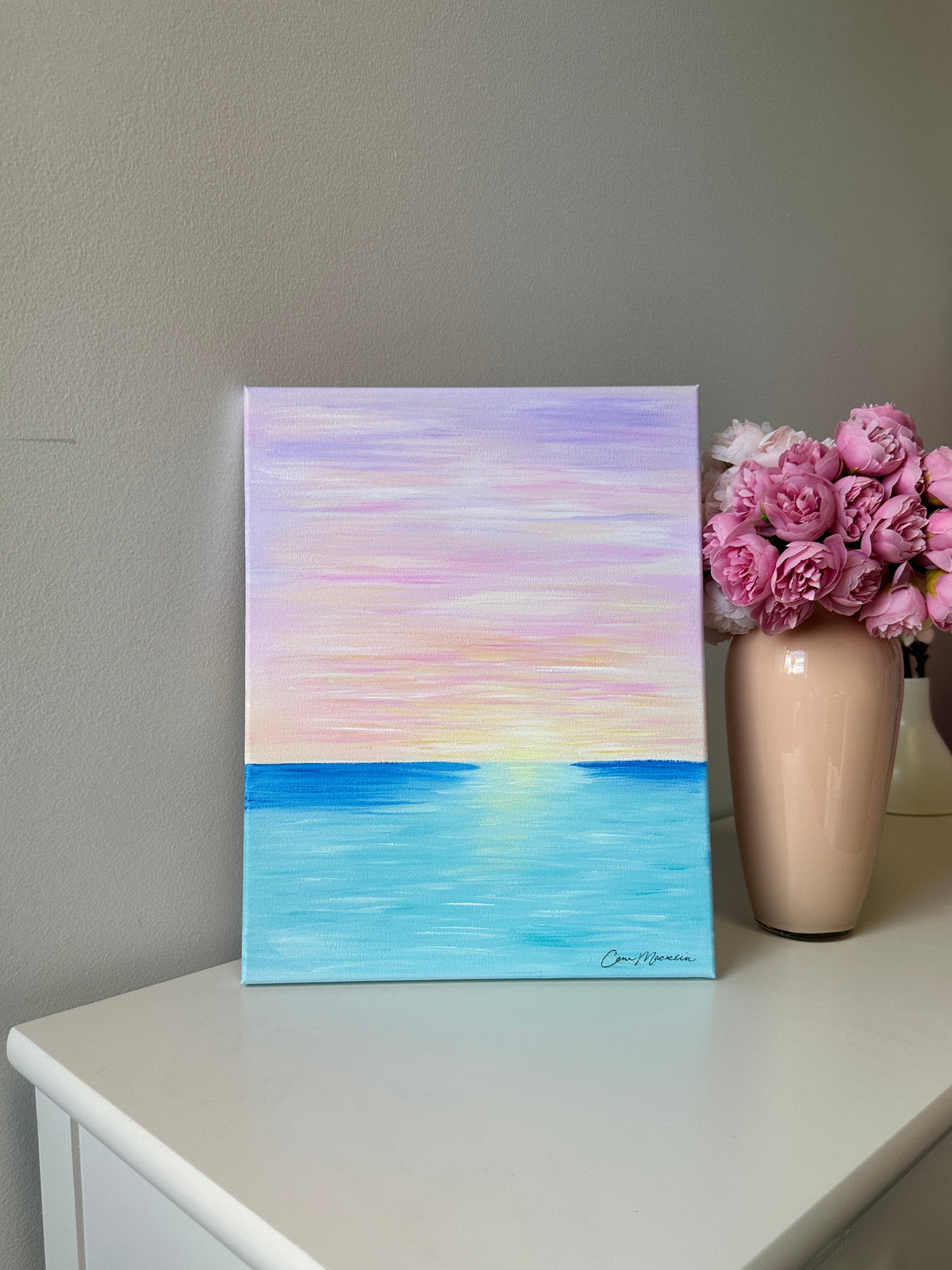 A square canvas painting of a pastel sunset over the ocean, displayed on a white dresser next to a vase of pink roses against a gray wall.