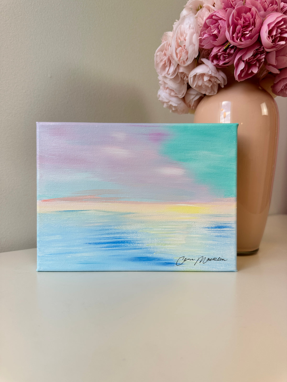 A small square canvas painting with an abstract pastel sky and ocean scene sits on a tabletop. The artwork features blended blue, aqua, lavender, pink, and soft yellow tones, with a visible artist signature in the lower right.