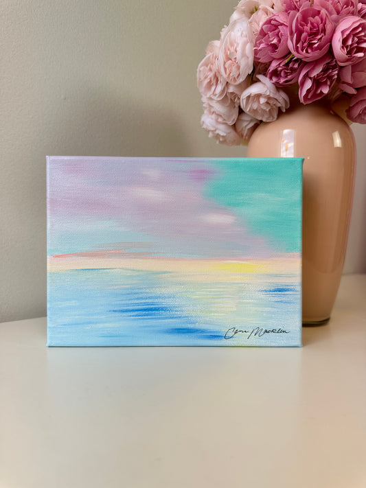 A small square canvas painting with an abstract pastel sky and ocean scene sits on a tabletop. The artwork features blended blue, aqua, lavender, pink, and soft yellow tones, with a visible artist signature in the lower right.