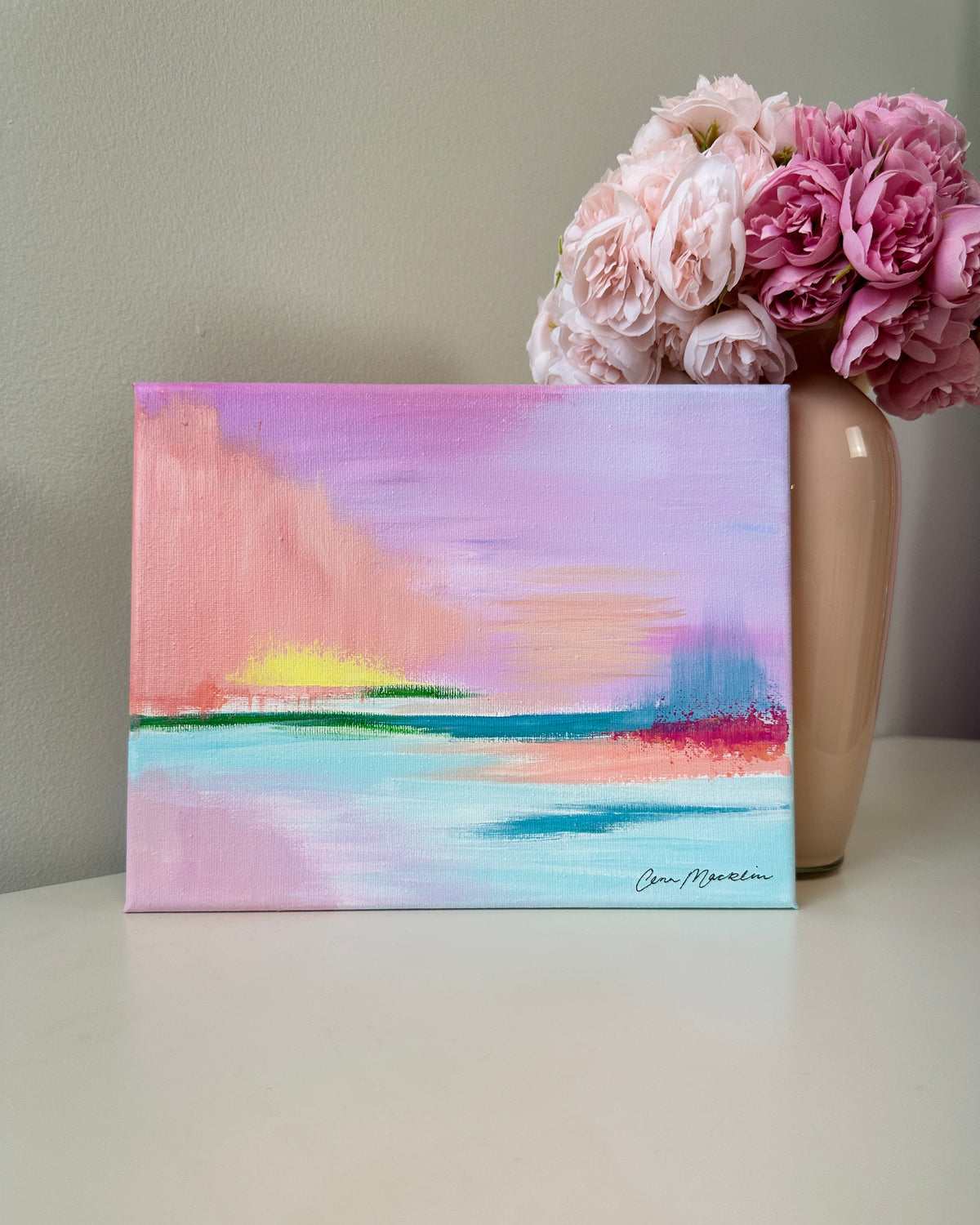 A square canvas painting with an abstract pastel landscape in pink, purple, blue, and peach tones, displayed on a white surface next to a vase of pink flowers.
