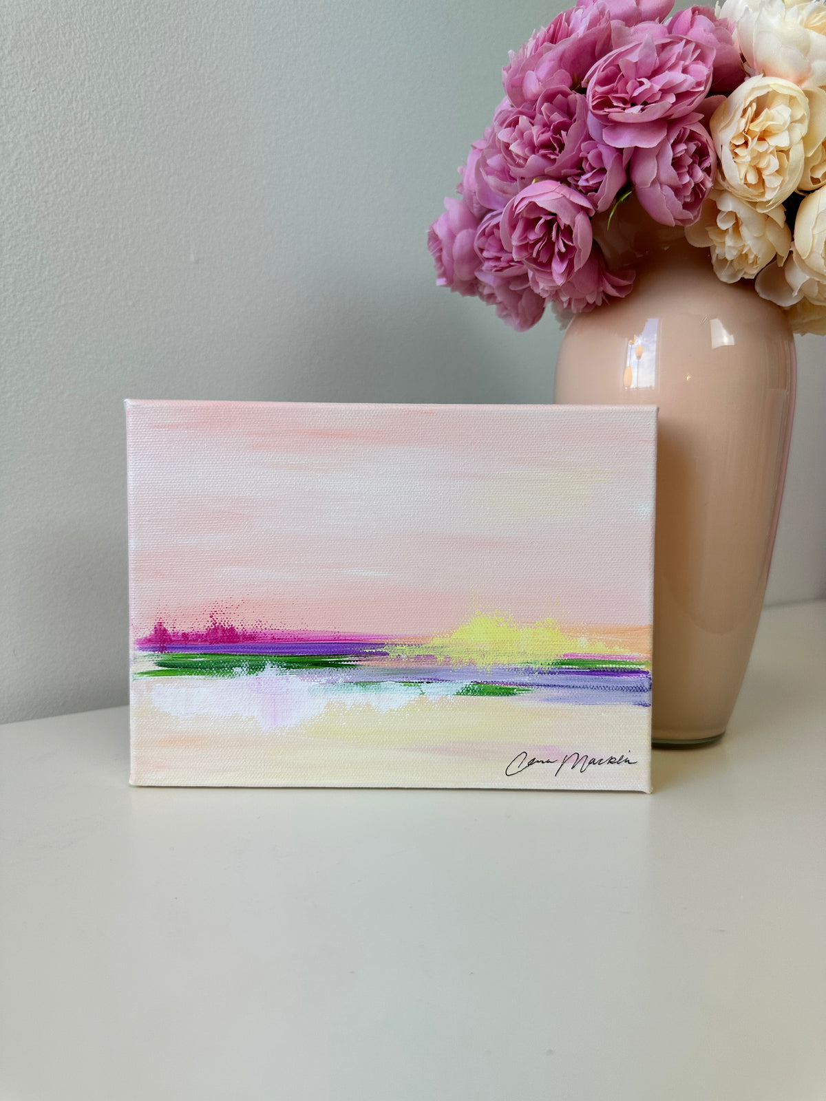 A small square canvas painting with an abstract landscape in pastel pink, purple, green, yellow, and white tones, displayed on a white surface next to a beige vase filled with pink and cream flowers.