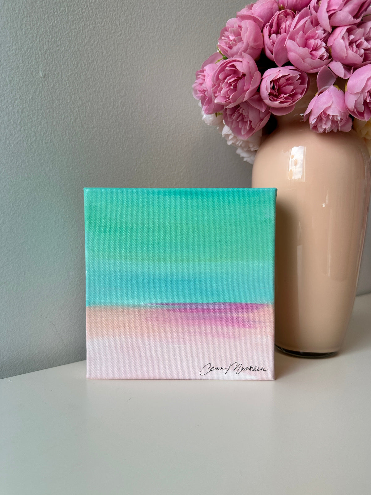 A small square canvas painting with a soft gradient design in teal, light blue, pink, and pale peach, displayed on a white surface next to a beige vase filled with pink flowers.