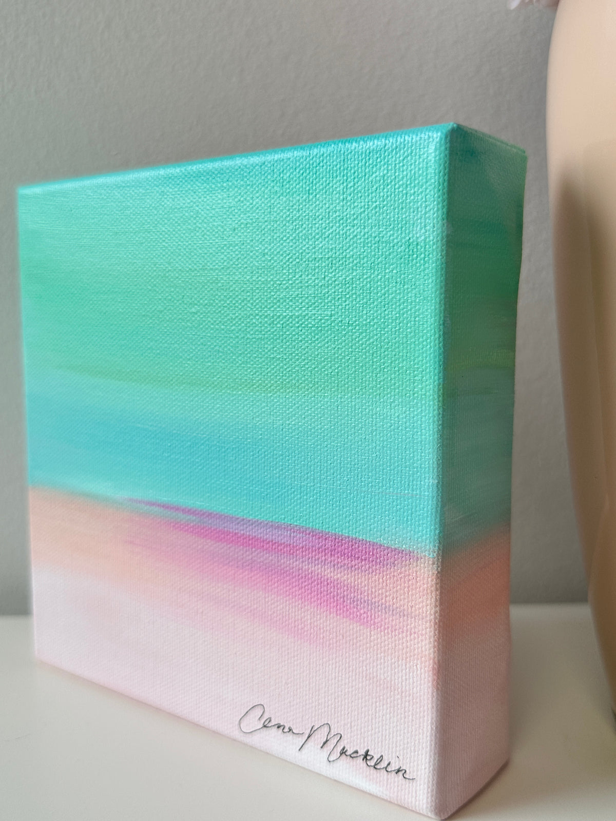 A small square canvas painting with a textured canvas finish, featuring a gradient design with a bright turquoise top half and a soft pink bottom half. The canvas is signed 'Cinn-McKeown' in black script at the bottom right corner, resting on a white surface against a neutral gray wall.