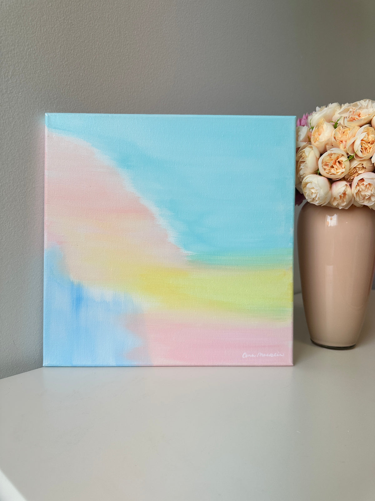 A square pastel abstract canvas painting with soft blue, pink, yellow, and white blended brushstrokes, displayed on a white surface next to a beige vase filled with peach roses against a gray wall.