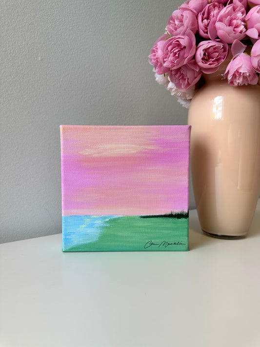 A small square canvas painting with a pink sky and green-blue landscape sits on a white surface next to a beige vase filled with pink flowers against a gray wall.