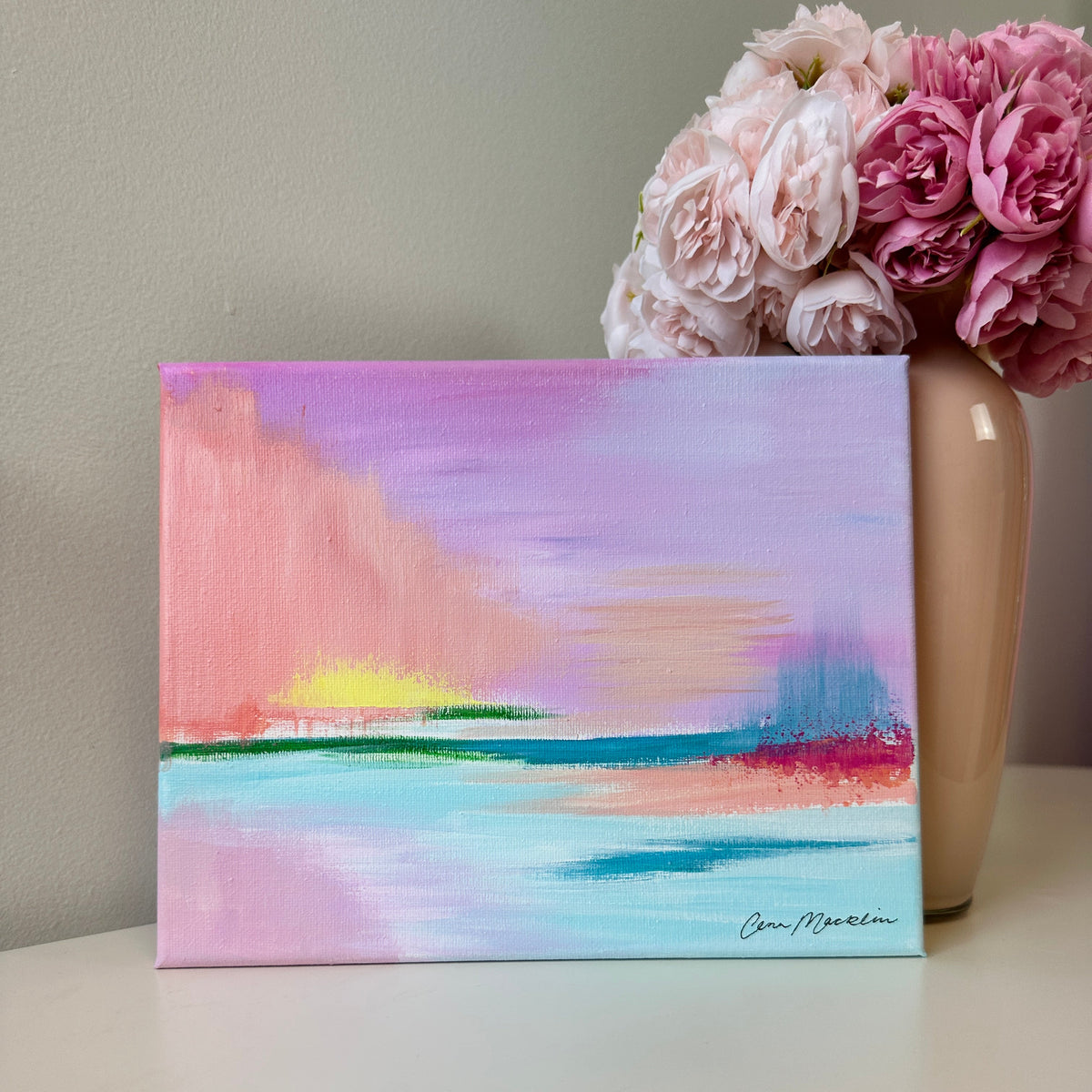 An abstract landscape painting on a canvas, featuring pastel shades of pink, purple, blue, and yellow, displayed on a white surface next to a beige vase with pink flowers.