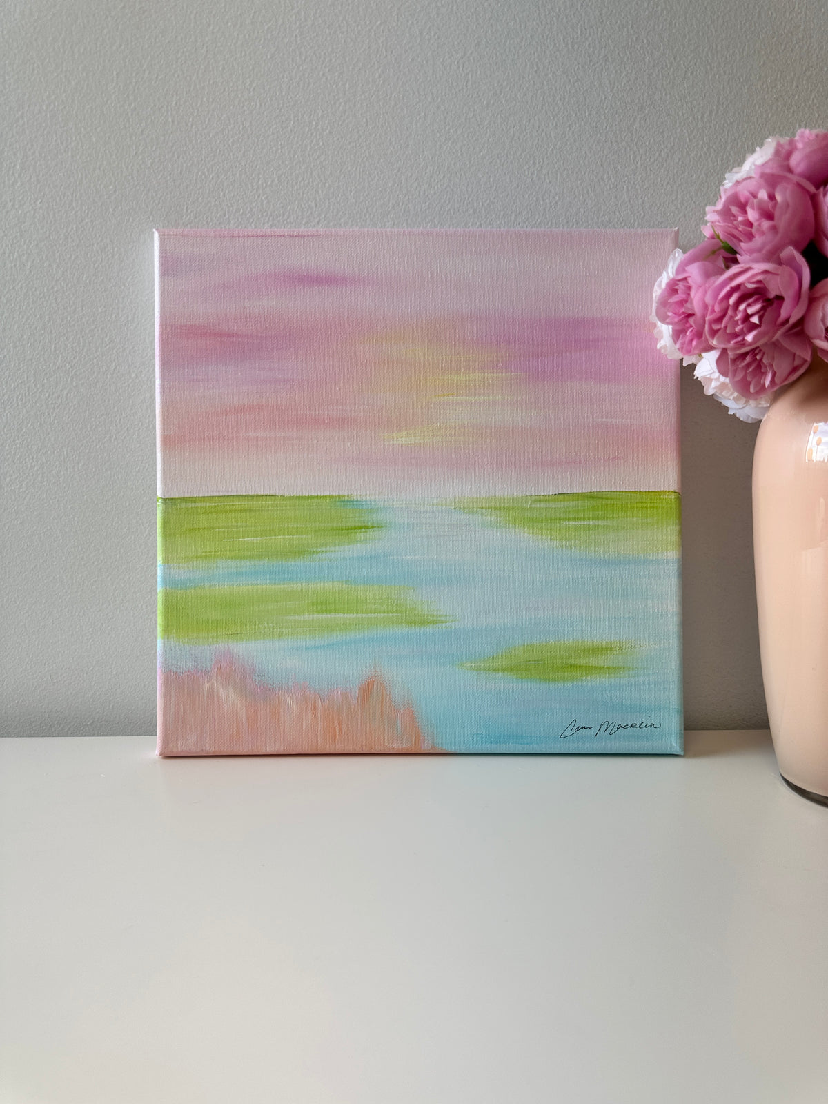 A square canvas painting with an abstract landscape design in pastel pink, green, blue, and peach tones, displayed on a white surface next to a vase of pink flowers against a gray wall.