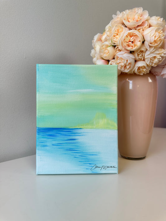 A small square canvas painting of a seascape with a green sky, blue water, and a distant yellow-green island, displayed on a white surface next to a beige vase filled with pale peach roses.