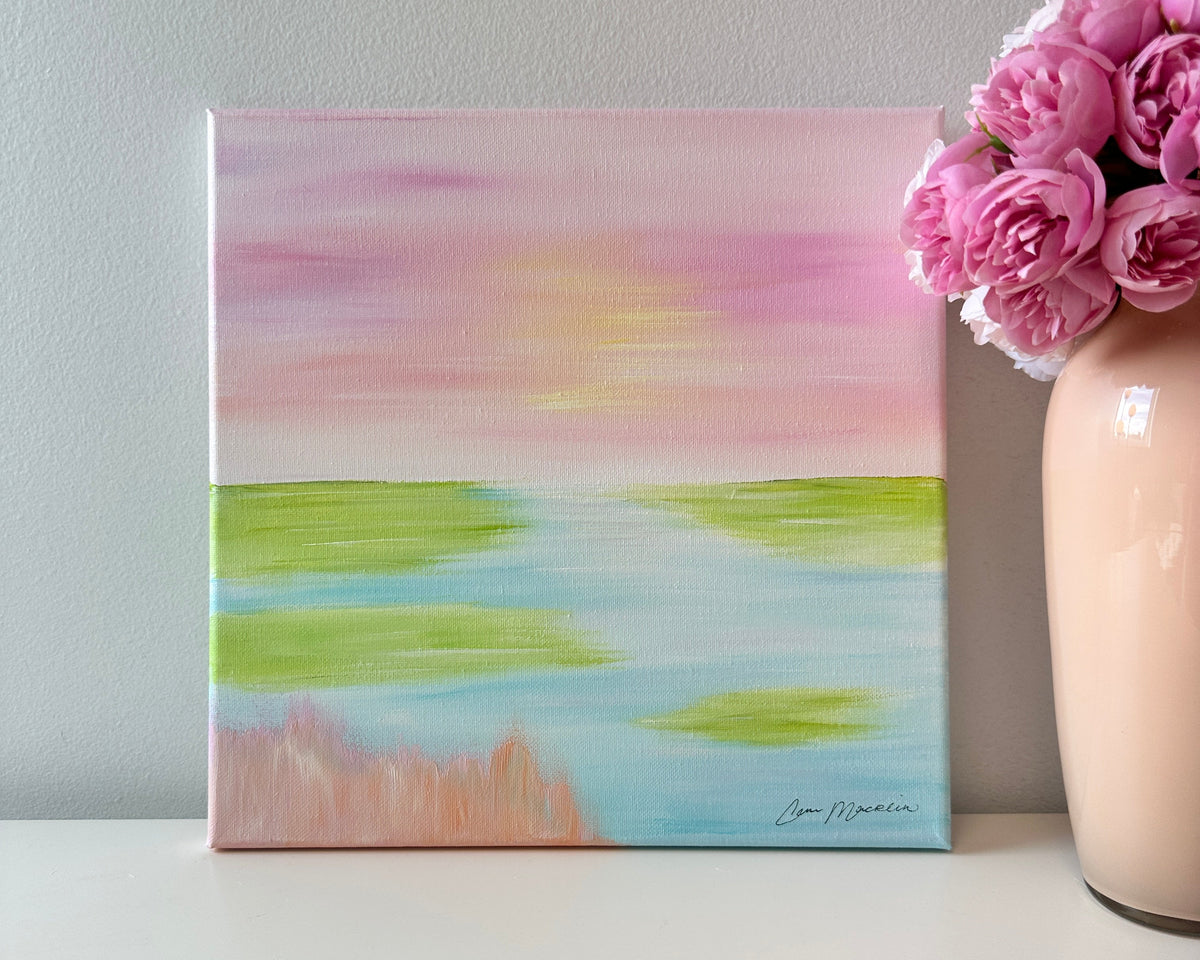 A small square pastel landscape painting with pink sky, green and blue water, and peach shoreline, displayed on a white surface next to a vase of pink flowers against a light gray wall.